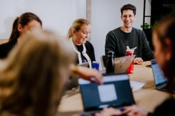 En gruppe kolleger sitter rundt et langt trebord i et kontormiljø, tilsynelatende i et møte. Flere har bærbare datamaskiner foran seg. En mann med briller og en mørk genser i bakgrunnen smiler, og en kvinne ved siden av ham ler. Andre personer er uskarpe i forgrunnen.