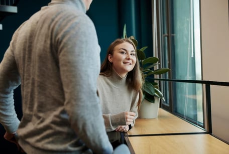 En ung kvinne i en beige høyhalset genser sitter ved en tre-disk ved et vindu. Hun smiler og ser opp på en person i en grå genser som står med ryggen til kameraet i forgrunnen.
