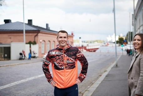 Smilende mann i oransje løpejakke med hendene i siden på en bygate, med en kvinne delvis synlig ved siden av.