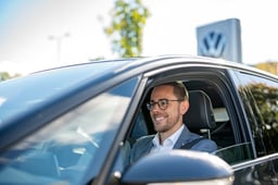 En mann med briller og dressjakke sitter i førersetet på en bil, smilende, med Volkswagen-logoen synlig i bakgrunnen.